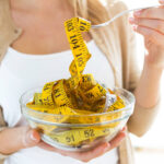 Worried cute woman holding bowl with measuring tapes at home.